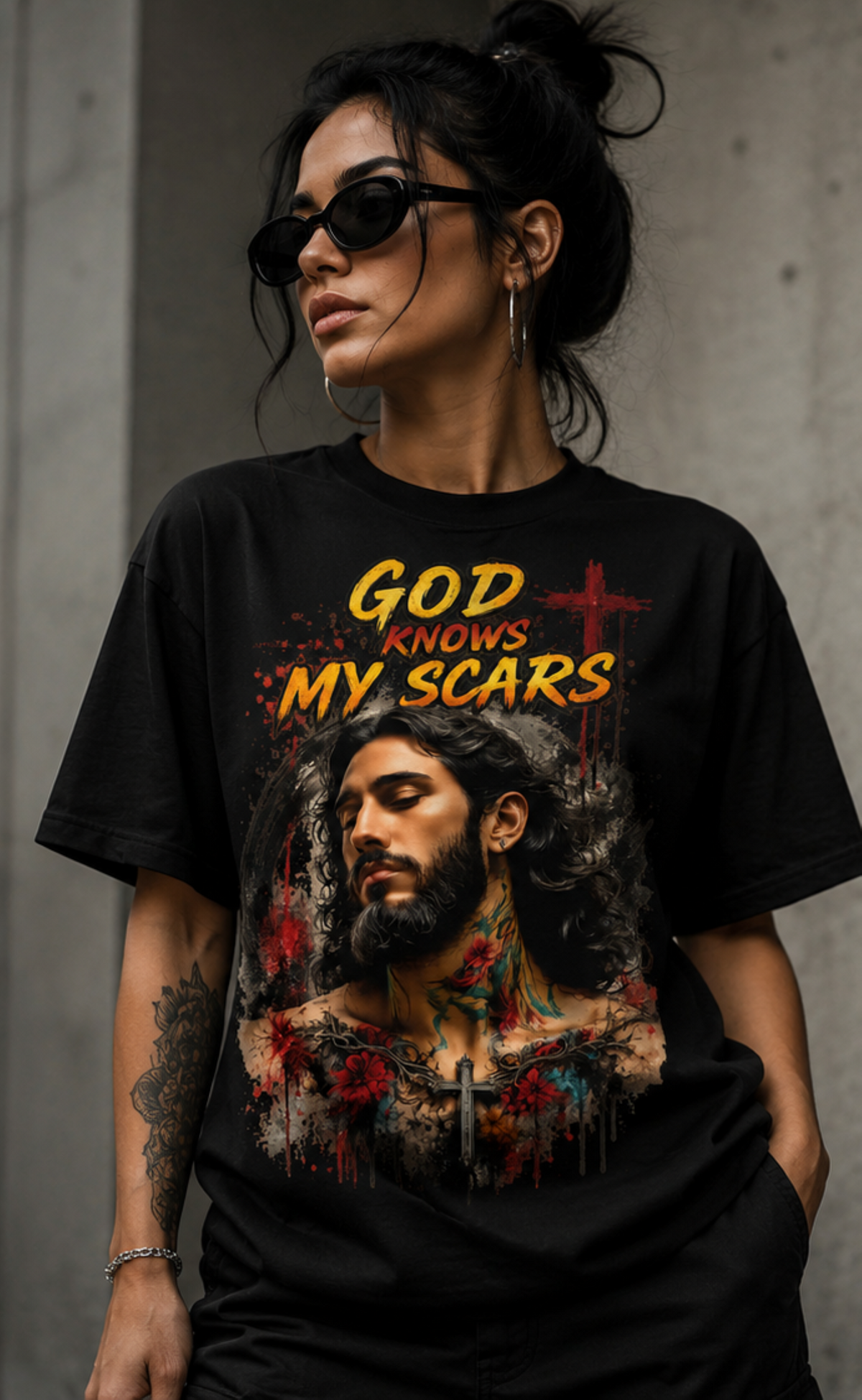 A woman wearing a black graphic t-shirt with the text 'GOD KNOWS MY SCARS' and a detailed portrait of Jesus with a cross, set against a concrete wall background.